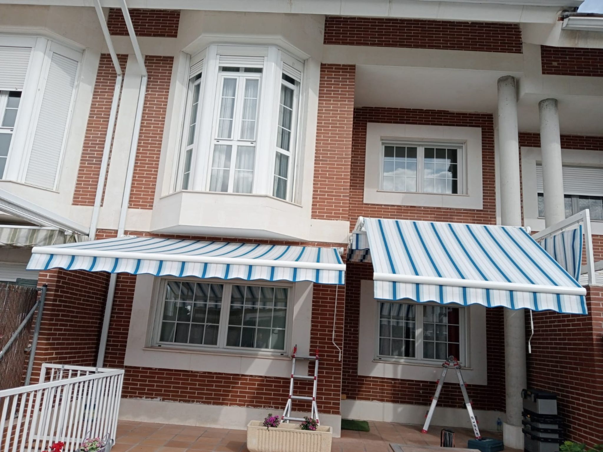 Toldos extensibles con rayas azules y blancas en fachada de vivienda unifamiliar de ladrillo y ventanas de mirador