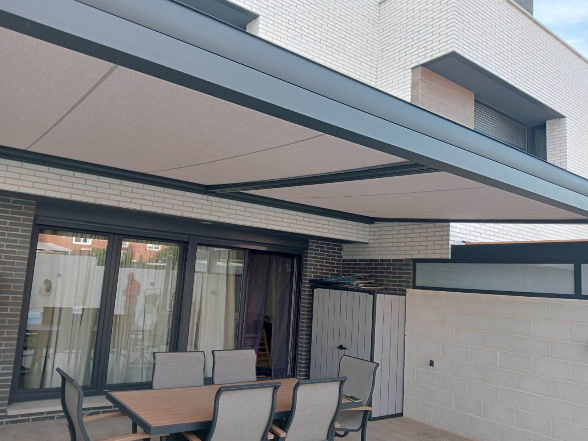 Pérgola moderna con estructura metálica y cubierta textil instalada sobre patio con mobiliario de comedor en vivienda adosada